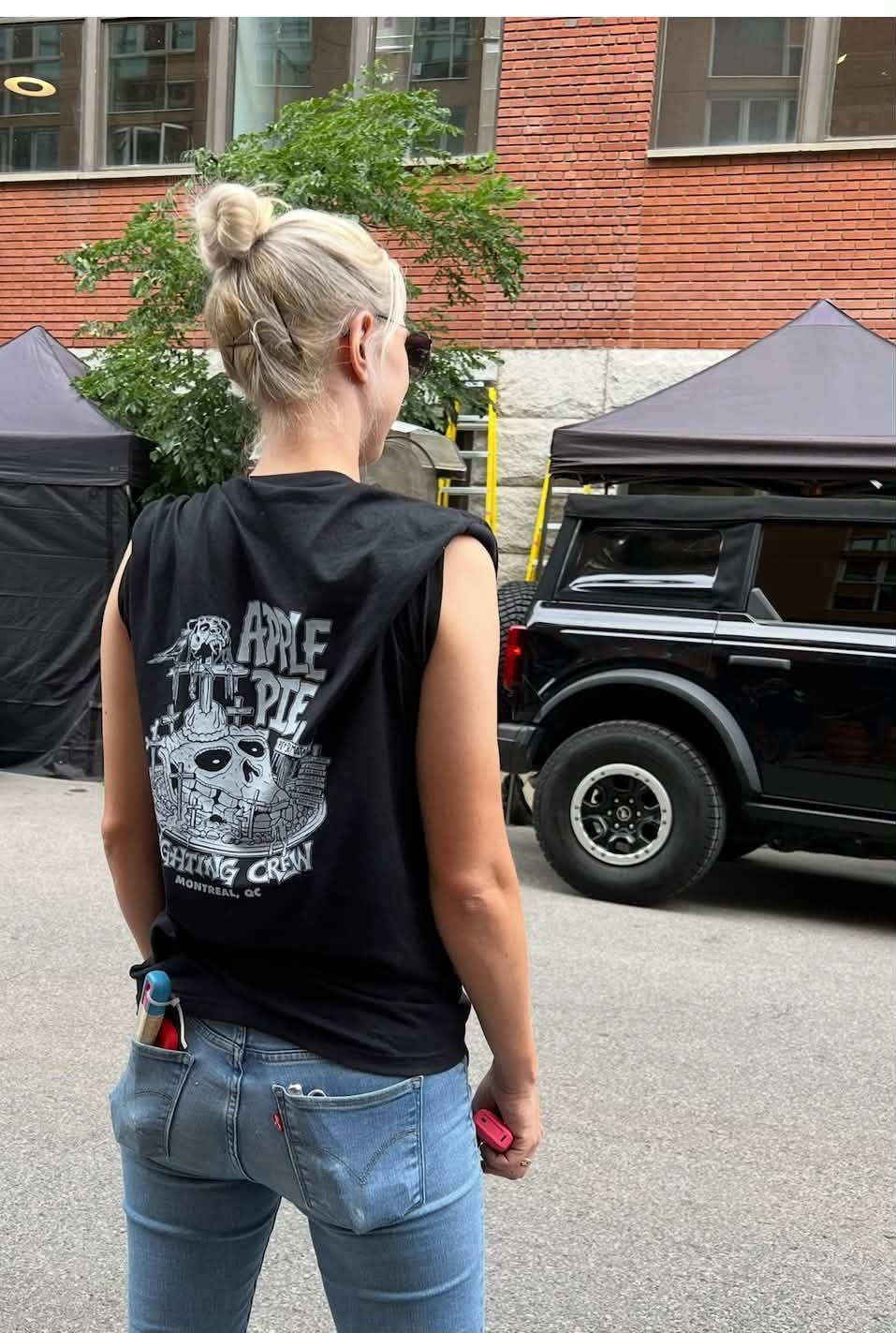 Technicienne d'éclairage vue de dos sur un plateau montréalais — tentes noires, Bronco de tournage, outils de gaffer dans la poche arrière — portant un t-shirt coupé « Apple Pie — Lighting Crew / Montreal, QC » imprimé par Dye Hard.
