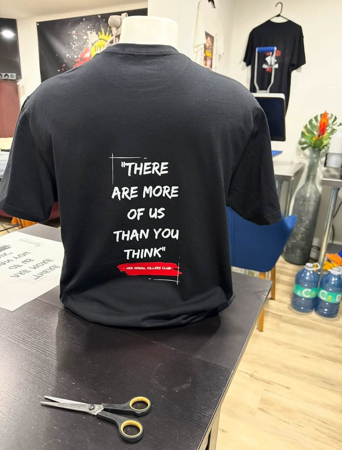 Dans l'atelier de Dye Hard : un t-shirt noir « There are more of us than you think — Her Serial Killers Club » sur un mannequin, ciseaux sur la table de travail, bannière au mur.